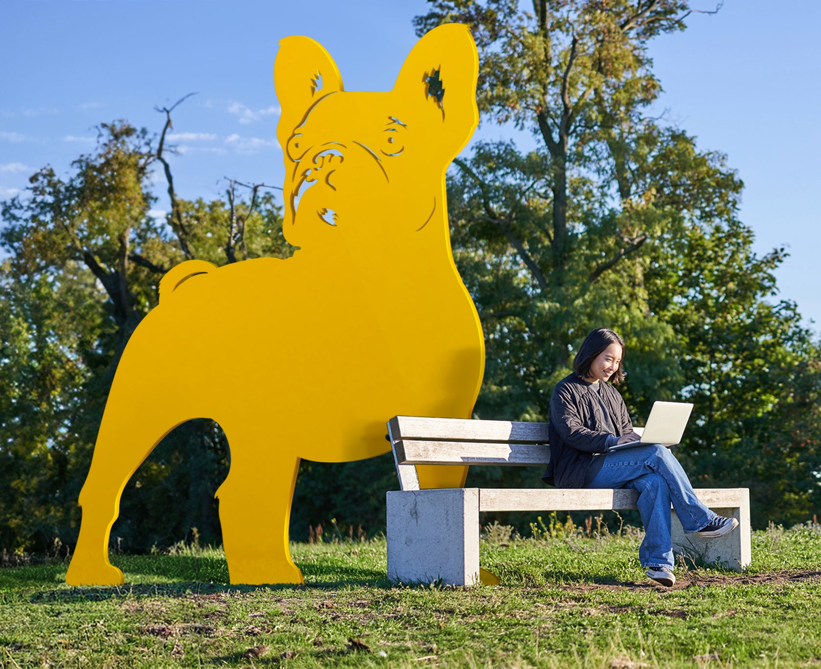 Statue d'un Bulldog de trois mètres de haut en métal laqué jaune dans un parc à côté d'un banc ou une jeune femme travaille.