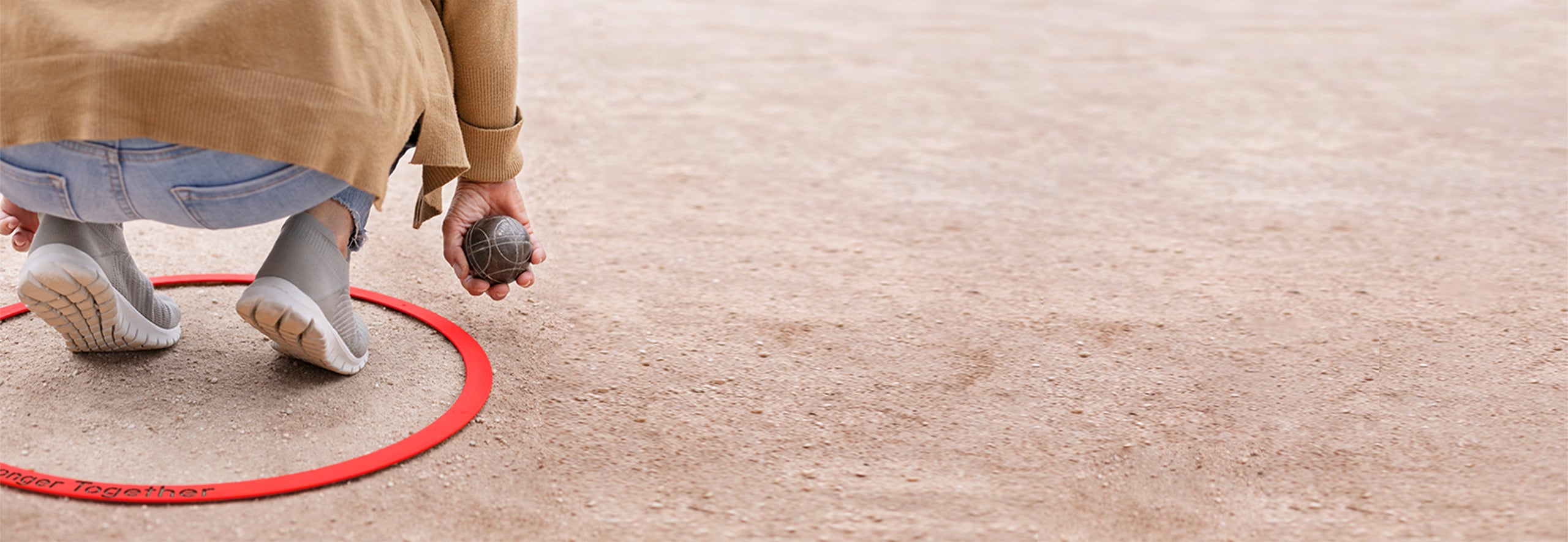 Gros plan sur un joueur de pétanque accroupi à l’intérieur d’un cercle en inox laqué rouge, tenant une boule prête à être lancée. Le cercle, posé sur le terrain, est gravé avec un message personnalisé.